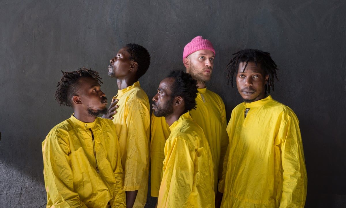 Five men, four black, one white dressed in yellow boiler suits stand in front of a grey wall