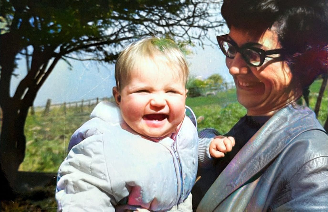 a smiling blonde baby wearing a pale anorak held by a smiling dark-haired woman with dark rimmed spectacles 