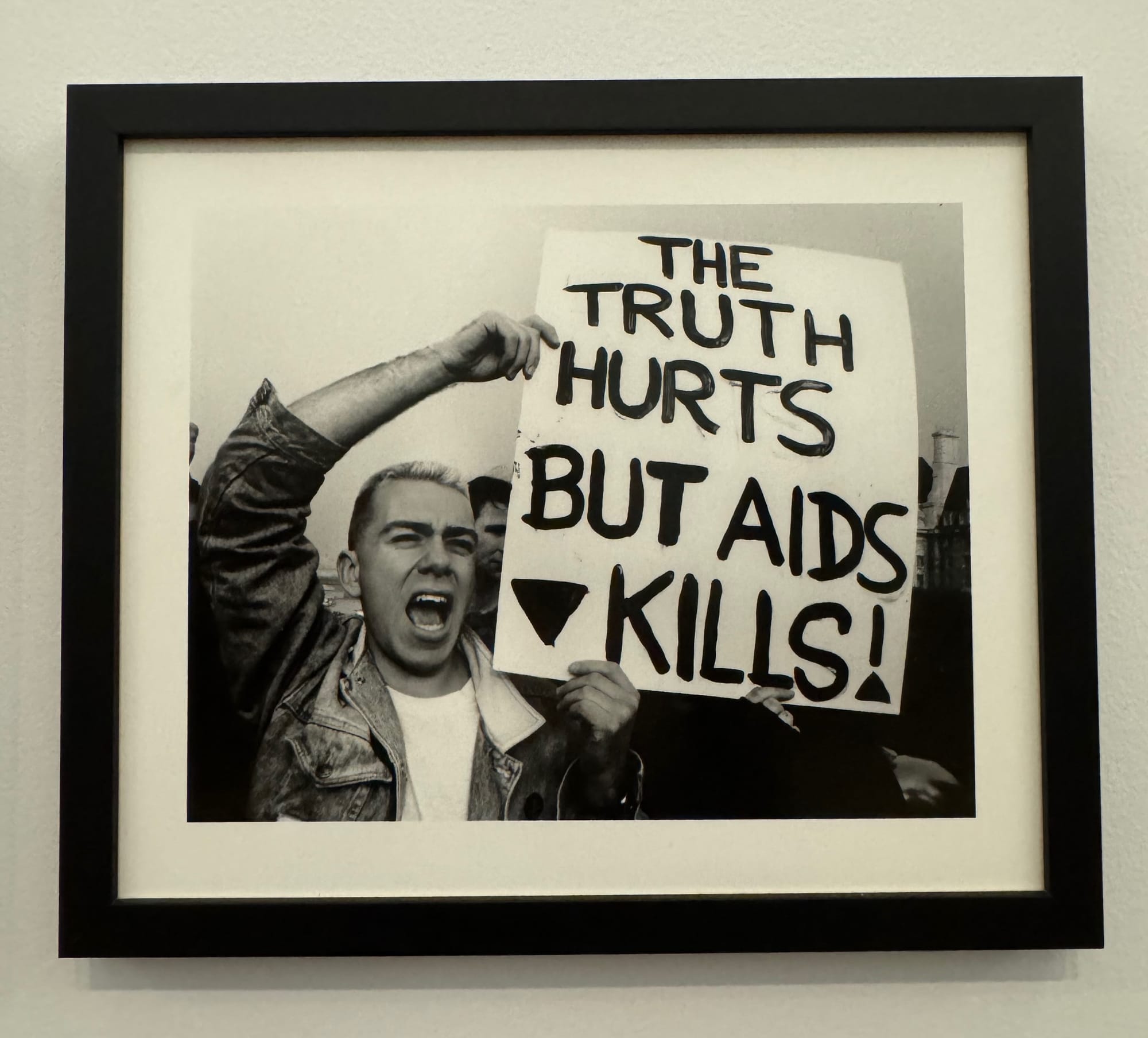 A black and white photograph in a black frame depicts a white man with his mouth open shouting. He is holding a placard which reads "The truth hurts but AIDS kills!"