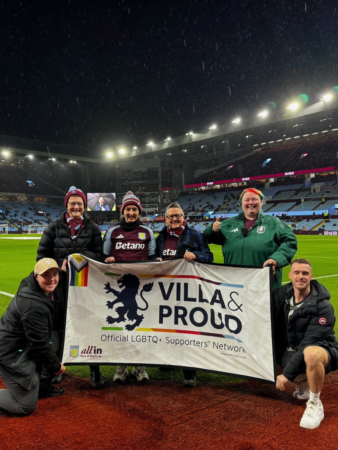 a group of people at a football ground hold up a banner which reads Villa & Proud official LGBTQ+ supporters' network