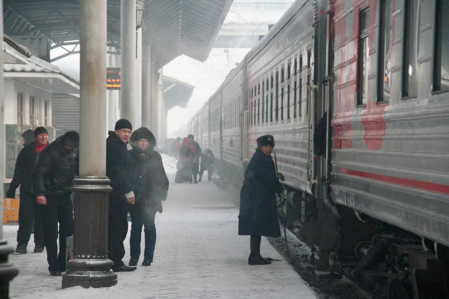 A grey and red train at a platform. A group in woolly hats and coats and scarves stand by a pillar. A guard is by the train