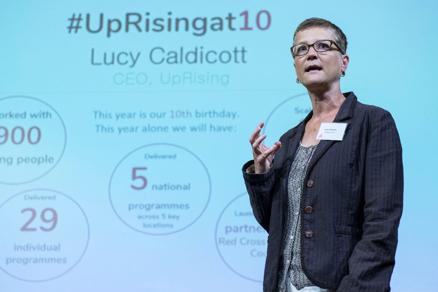 A woman in a brown jacket wearing glasses stands in front of a slide which reads: #UpRising at 10