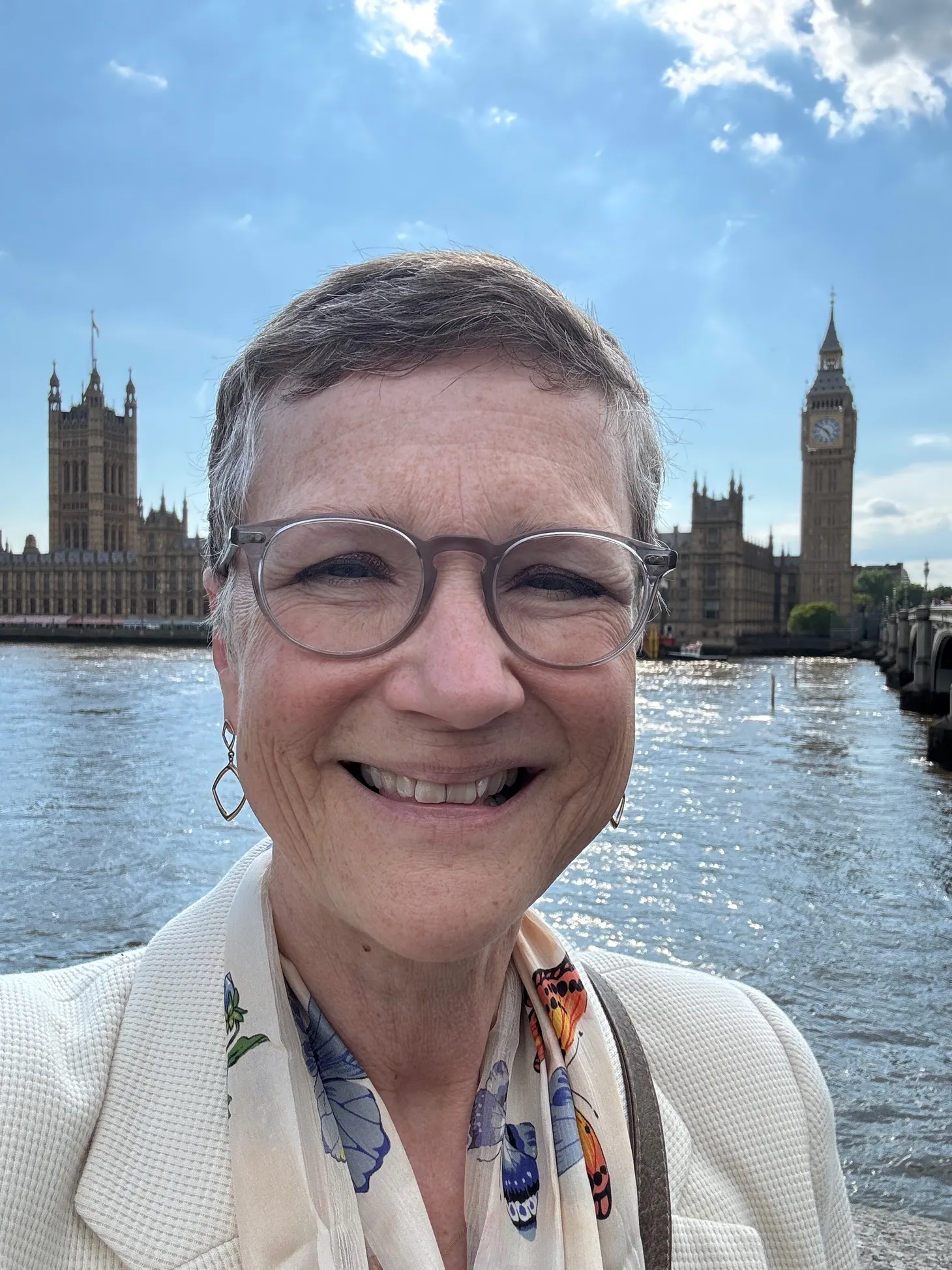 A white woman with short hair and glasses stands by a river with historic buildings on the opposite bank